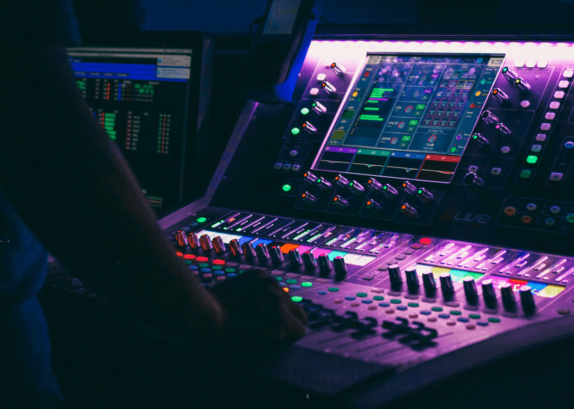 A sound and lighting control console glowing purple and blue, with faders, knobs, and a touchscreen interface being operated by a person in a dark room.